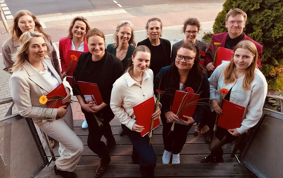 Zu sehen sind elf Personen, die gemeinsam auf der Treppe der Offenen Mühle Neubrandenburg posieren. Die Absolventinnen und Absolventen unter ihnen halten rote Zertifikatsmappen in die Hand und lächeln. 