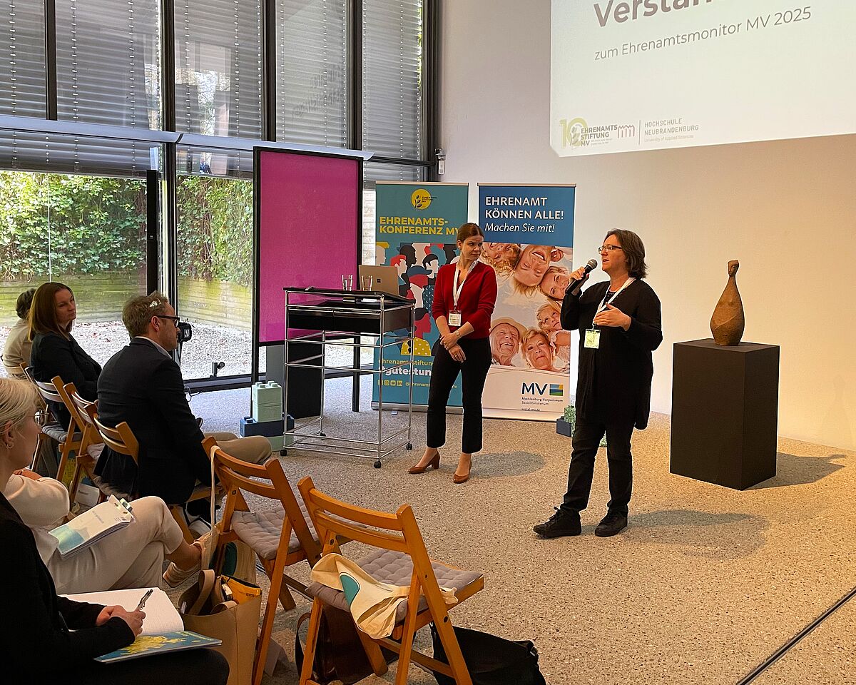 Zu sehen sind Professorin Christine Krüger und Professorin Claudia Vogel bei der Präsentation des Ehrenamtsmonitors 2025. Sie reden vor sitzendem Publikum. Hinter ihnen befinden sich zwei Rollups zur Ehrenamtskonferenz. An der Wand ist eine Projektion einer Powerpoint-Präsentation zu erkennen.
