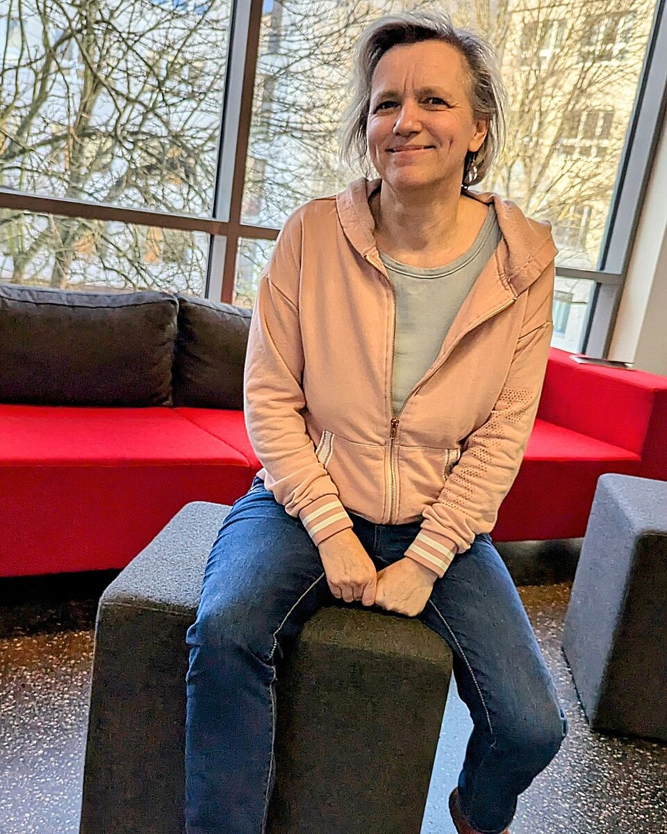 Das Foto zeigt Kathrin Minstedt, sitzend auf einem grauen Würfel, im oberen Foyer des Hochschulhaupthauses. Im Hintergrund befindet sich ein rotes Sofa. Der Innenhof des Hauses ist durch große Fensterfronten zu erkennen.