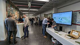 Das obere Foyer von Haus 1 ist gefüllt mit Menschen an Stehtischen und Informationsständen. Zu sehen ist  auch ein großer Monitor, welcher eine Karte mit der Überschrift "All my fields" zeigt. Im Hintergrund melden sich Personen am Einlass vor Hörsaal 1 für das Ergebnis-Symposium an.