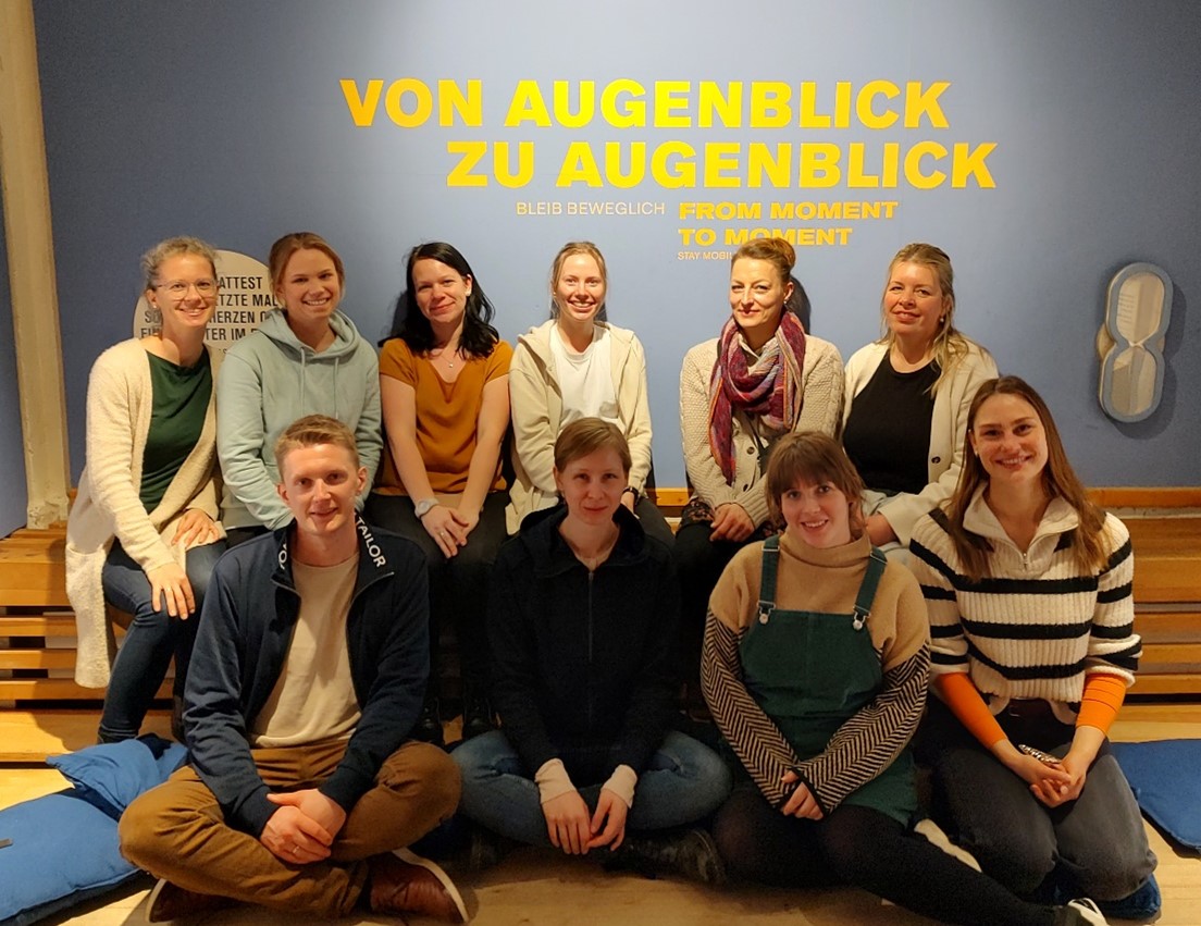 Auf dem Foto sind Prof. Dr. Katja Zehbe und 9 Studierende des Studiengangs "Pädagogik der Kindheit (berufsbegleitende)" zu sehen. An der Wand hinter Ihnen steht der Spruch "Von Augenblick zu Augenblick"